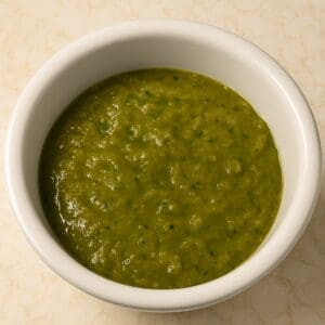 A bowl of roasted tomatillo and serrano salsa surrounded by fresh ingredients like tomatillos, serrano peppers, onion, and garlic.